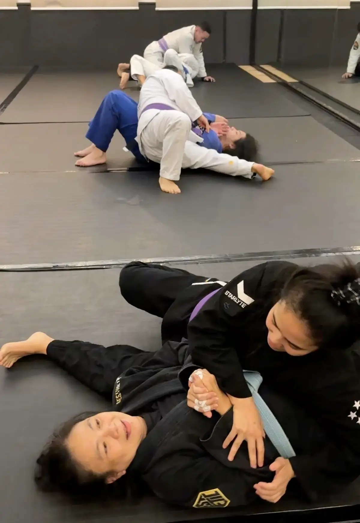 Two VCBJJ female members drilling in black gis, both smiling mid-roll — the relaxed energy shows the non-intimidating training environment