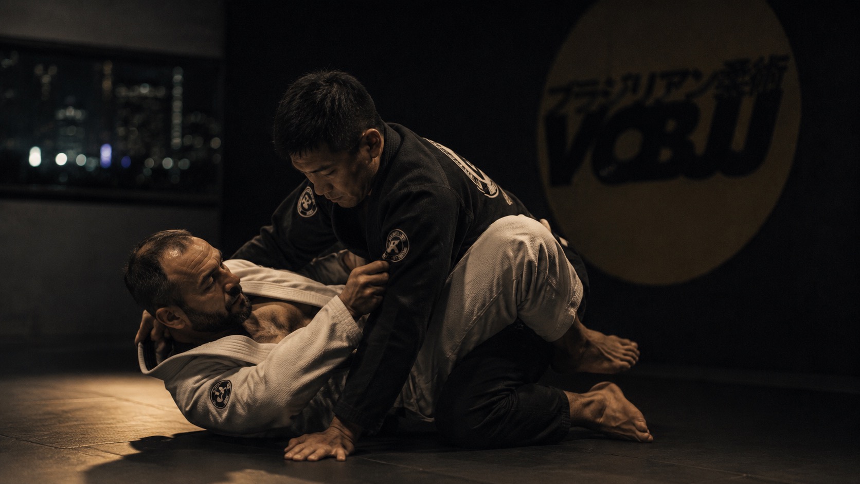 A training room in Kuala Lumpur — mats lit by a single overhead source, two figures mid-exchange, the city visible as a blur through a narrow window