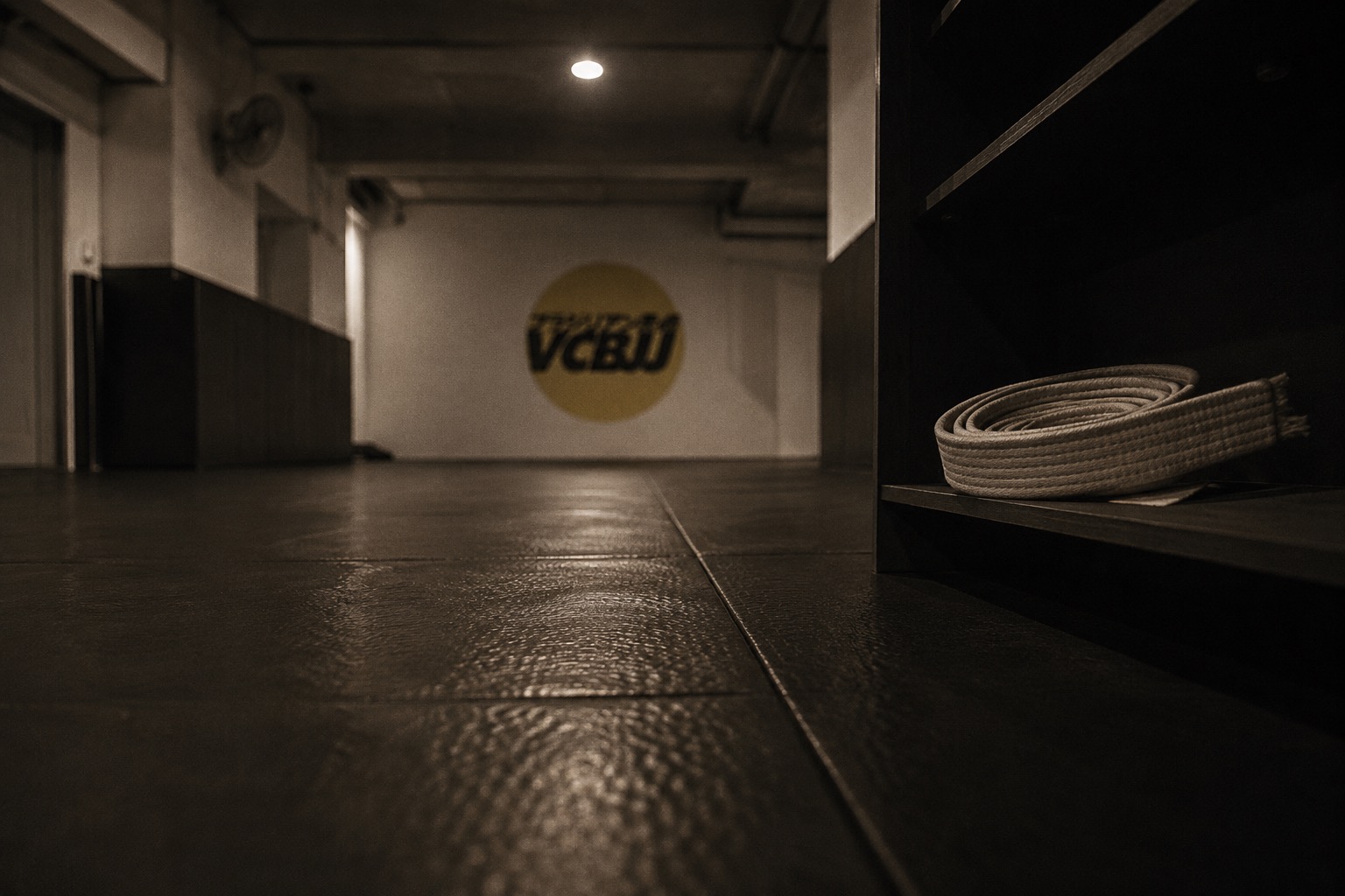 The VCBJJ training room in Bangsar — mats empty, a white belt coiled on a shelf in the foreground, the logo on the far wall